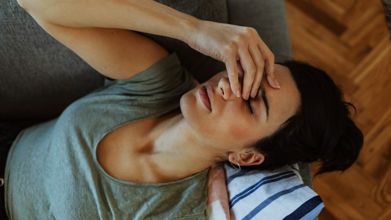 Unhappy Woman Touching Her Head and Suffering From Headache. Portrait of an Attractive Young Woman Sitting on a Sofa at Home With a Headache, Feeling Pain and With an Expression of Being Unwell. People, Emotions, Stress and Health Care Concept