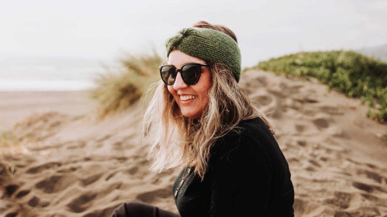 Smiling blonde plus size woman dressed in black, wearing a headband, sunglasses and sitting on the beach. Overcoming depression